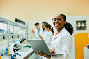 Happy black biochemist working on laptop in lab and looking at camera.