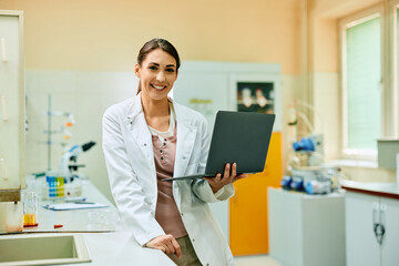 Happy female scientist working on laptop in laboratory and looking at camera.
