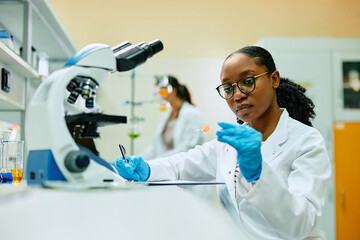 Young black biochemist examining sample on microscope slide during her research in lab.