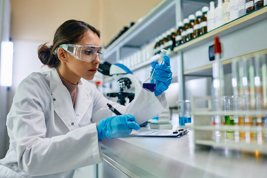 Female scientist taking notes while analyzing blue chemical liquid in lab.