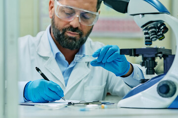 Scientist taking notes while analyzing microscope slide during research in lab.