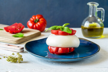 Burrata buffalo cheese with tomatoes, fresh basil, olive oil and dry oregano on a blue plate over white wooden table with ingredients and dark blue background. Close up