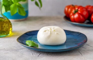 Burrata buffalo cheese on a blue plate, red tomatoes, olive oil, fresh basil over concrete background. Close up. Caprese salad ingedients