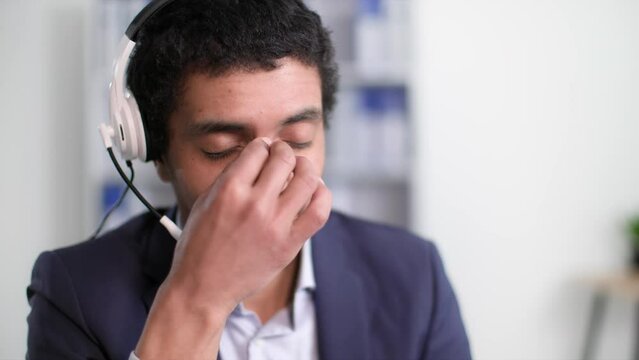 hard work, tired african american man takes off headset and eyeglasses due to fatigue and rubs eyes with hands after working day sitting at table in office