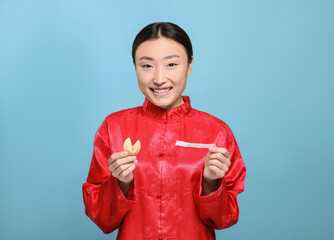Asian woman holding tasty fortune cookie with prediction on light blue background, space for text