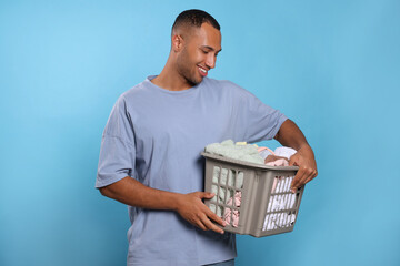 Happy man with basket full of laundry on light blue background