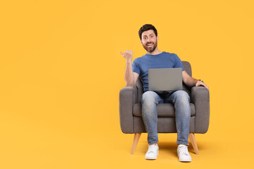 Emotional man with laptop sitting in armchair against yellow background. Space for text
