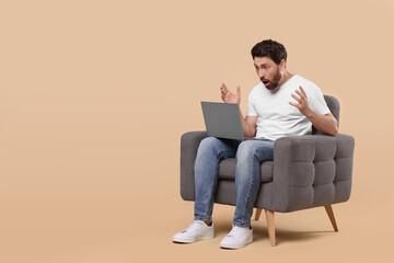 Emotional man with laptop sitting in armchair against beige background. Space for text