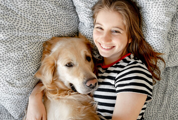 Pretty girl hugging golden retriever dog and smiling lying in bed. Happy teenager with purebred doggy pet labrador resting together