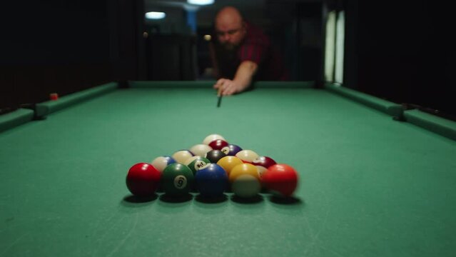 Bald bearded man breaks the pyramid of balls by hitting the white cue ball with the cue close up. Billiard player starts the game with his first shot