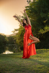 brunette woman in an elegant red party dress on the green grass of a beautiful natural landscape with a lake at sunset, with her arms raised and showing her toned dancer's leg out of the dress. 