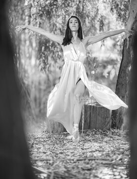 black and white photo of an attractive brunette girl in the forest, framed between 2 trees, smiling and with her arms outstretched, wearing an open dress revealing her leg and ballerina shoes.