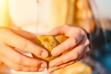 Woman's hands opening a fig to eat. Fresh ripe fig fruit in the hands. A young woman eats figs on the beach, near sea. Ficus carica, Fig - green-yellow fruit. Close up. Selective focus