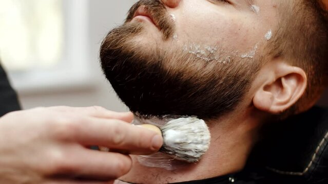 Hairdresser barber master applies shaving foam to face of male client sitting in barbershop chair, closeup. Man with beard sits in chair while barber applies shaving cream