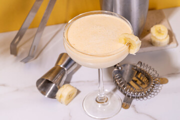  Sweet summer cold banana daiquiri or martini cocktail, liquor with banana syrup, slices,crushed ice and bar utensils on high-colored golden beige background 