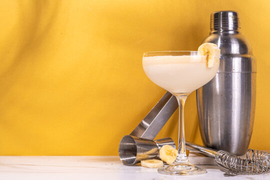  Sweet summer cold banana daiquiri or martini cocktail, liquor with banana syrup, slices,crushed ice and bar utensils on high-colored golden beige background 