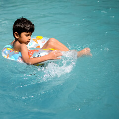 Fototapeta na wymiar Happy Indian boy swimming in a pool, Kid wearing swimming costume along with air tube during hot summer vacations, Children boy in big swimming pool.