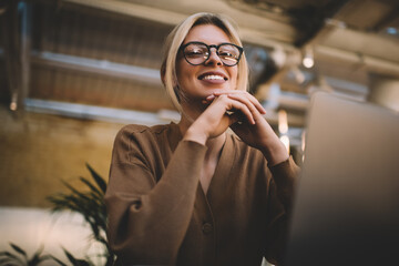 Portrait of cheerful Caucasian female with blurred laptop smiling at camera during time for web networking indoors, happy millennial blogger in optical spectacles for vision protection posing