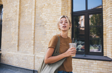 Pensive Caucasian tourist with takeaway ice coffee exploring new city during summertime vacation journey for recreation, contemplative hipster girl with caffeine beverage walking around town