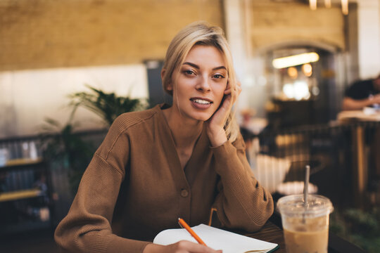Caucasian female student looking at camera and studying coffee time in cafe interior, portrait of young blonde woman with education notebook for learning doing homework and posing indoors