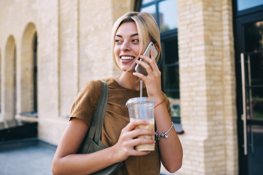 Happy female user with ice coffee to go making roaming communication via cellular application, cheerful millennial hipster girl calling via mobile technology for discussing travel vacations