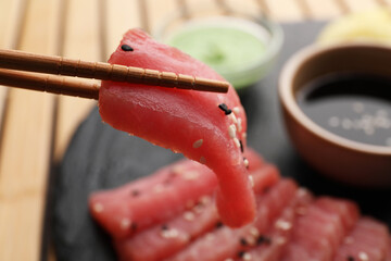 Holding tasty sashimi (piece of fresh raw tuna) with chopsticks against blurred background, closeup