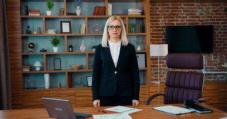 Fototapeta na wymiar Adorable confident experienced blond businesswoman in business suit posing on camera near her workplace in well-designeted office room