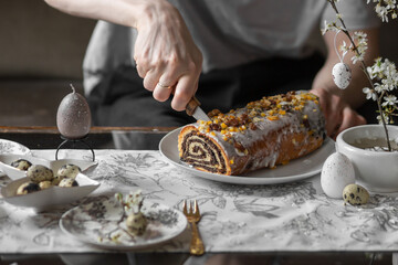 Obraz na płótnie Canvas Man cutting poppy seed roll with knife. Festive table with poppy seed Easter cake, quail eggs, candles, spring branches.