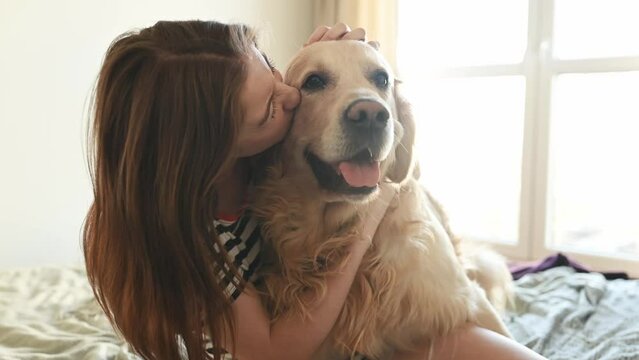 Pretty girl hugging golden retriever dog and smiling sitting in bed. Female teenager with purebred doggy pet labrador looking at camera at home