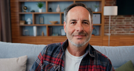 Portrait of good-looking joyful relaxed adult bearded man in checkered shirt,white t-shirt and jeans which dreamly looking to window in living-room then turning his head into camera