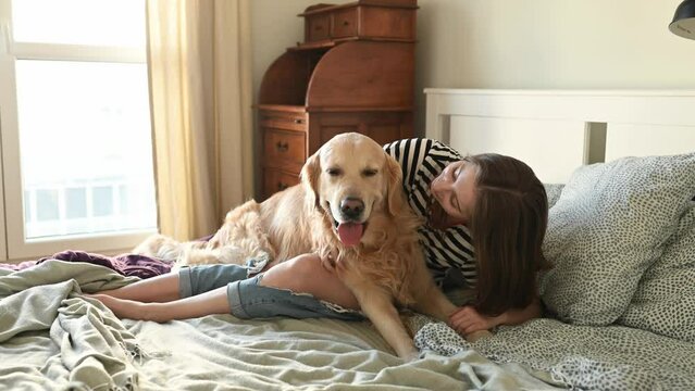 Pretty girl hugging golden retriever dog and smiling sitting in bed. Female teenager with purebred doggy pet labrador looking at camera at home