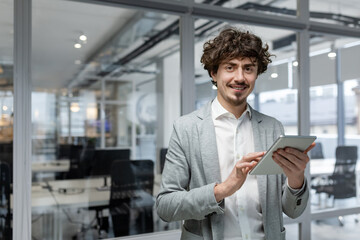 Naklejka na ściany i meble Portrait of successful young businessman, man with tablet computer in hands smiling and looking at camera, entrepreneur at workplace inside modern office.