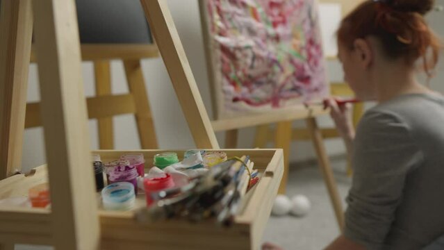 The artist creates a drawing on an easel using bright paints and brushes in the studio. Artist's hands of young woman reflect her creation process as she creates her own masterpiece painting on easel