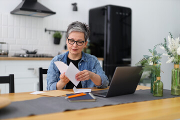 Serious mature woman at a table in a home interior looks through mail and bank accounts.