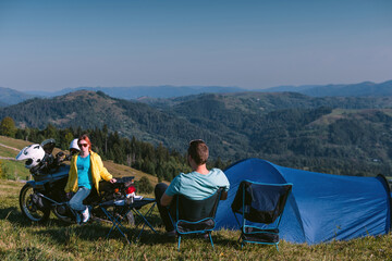 A motorcyclists, man and woman. Rest with a deep view. Loving couple of tourists. Relationship and adventure concept. motorcycle, tent. Active lifestyle. Folding chairs. Top mountains.