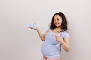 Attractive positive young pregnant woman with big belly, pointing finger at blue knitted baby booties on her hand, smiling with a cheerful toothy smile, isolated on white background. Copy ad space