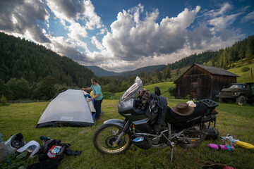 Setting up a blue tent with a great view of the mountains. A trip on a motorbike. A touring motorcycle with lots of gear. Bags, luggage. A couple of bikers, stopped for the night in a camp.