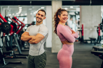 A cheerful confident sporty couple is standing in a gym with arms crossed and smiling at the camera.