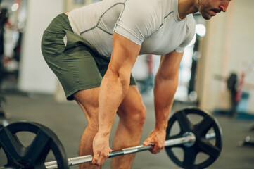 Cropped picture of a muscular sportsman bending and lifting a barbell in a gym.