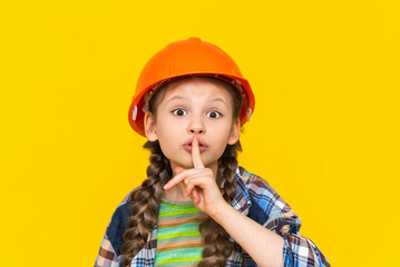 A happy little girl is wearing a construction helmet and a shirt, pointing her finger to be quiet. The child chooses repairs in the nursery. Yellow isolated background.