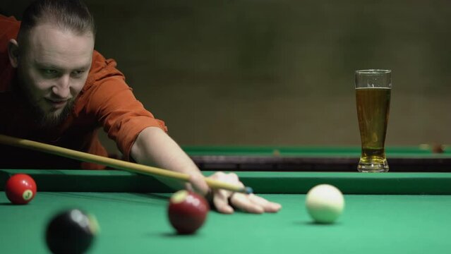 Man drinking beer and playing billiard with cue and balls in a green pool table. Active leisure and entertainment