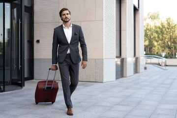 Smiling and good looking manager with suitcase walking outside modern office.