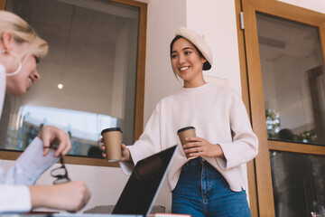 Happy Asian freelancer in trendy beret giving takeaway coffee cup to blurred colleague enjoying cooperation meeting in coworking, joyful hipster girls talking and smiling near table with netbook