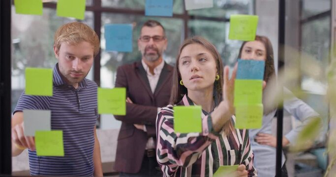 Team of caucasian business people standing near glass window with sticky notes. Scrum methodology with colorful stickers. Work colleagues discussing project ideas.