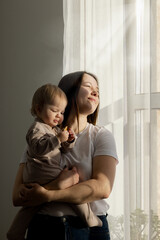 Young beautiful mother holds her baby in her arms, looks out the window and squints in the sun