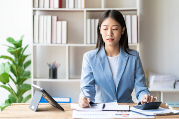 Asian female accountant holding pen working on financial accounting and calculator to calculate business data, accounting documents and laptop computer at office