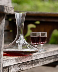 Decanter and two glasses with red wine on wooden table. Glass smooth transparent decanter. Aged gray wood and blurred greenery background. Wine tasting. Side view. Copy space. Soft focus. 