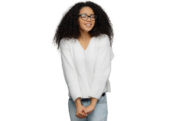 Happy woman with frizzy hair and glasses, wearing white jumper and jeans, keeps hands together, smiles with eyes shut, isolated on grey background. Good emotions concept