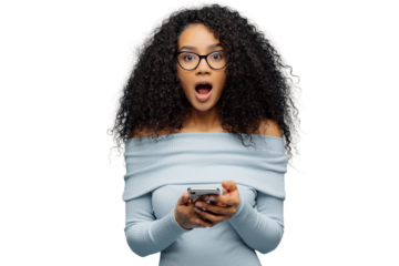 Amazed woman with Afro hair reads phone, shocked by online post, wears glasses and sweater, isolated on white background