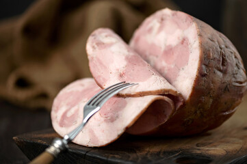 Piece of ham is cut into slices on wooden board. End of sausage. Slice of ham on fork. Dark...